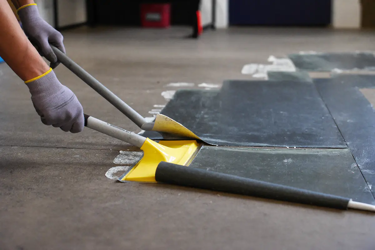 Handen in werkhandschoenen die met een lange vloerschraper verlijmde vinylstroken van de vloer losmaken